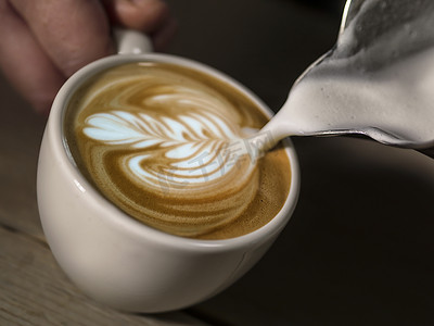 拿杯子摄影照片_“亲手制作拿铁咖啡馆的咖啡师，白杯上带有精美拿铁艺术的咖啡”