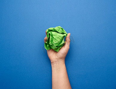 蓝色褶皱纸摄影照片_蓝色背景上拿着皱巴巴的绿纸球的女手