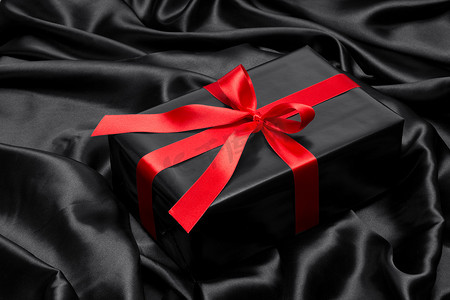 “带红色缎带和蝴蝶结的黑色礼盒，覆盖在黑色缎面上”
