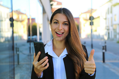 西班牙女摄影照片_拿着智能手机的相当美丽的年轻女商人在街上竖起大拇指。