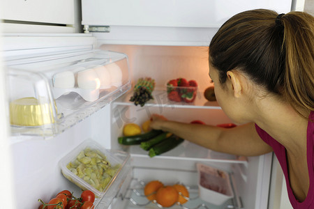 冰箱里的水果摄影照片_在冰箱里寻找健康食物的年轻女性的后视图