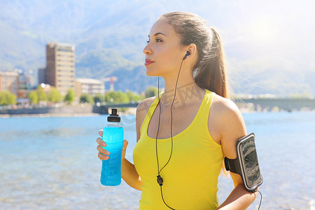 口渴的女运动员带着智能手机的臂章拿着能量饮料，在户外看着别处。