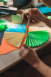 在前景中，女性手在木桌上粘着折纸扇、彩色纸片和剪刀。