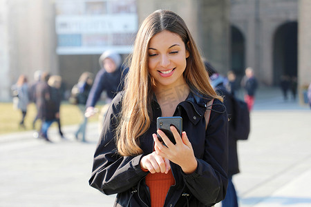 阳光明媚的春日，带智能手机的快乐美丽年轻高中女生在户外发短信和微笑