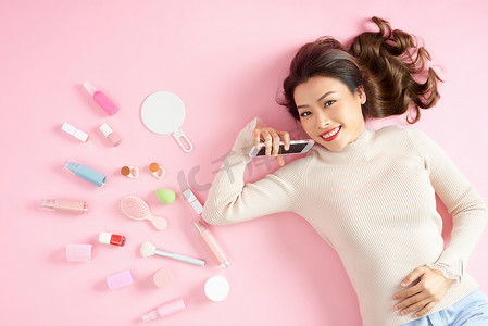 美女俯视图摄影照片_年轻的亚洲美女用她的化妆工具展示智能手机 — 她躺在粉红色的地板上。