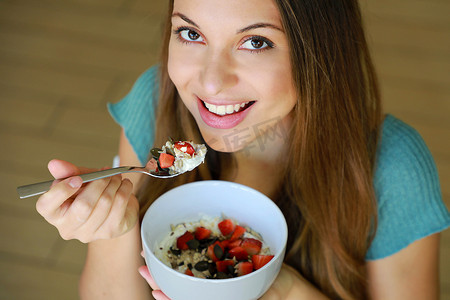 从上方特写美丽的年轻女子微笑着在家里吃着麦片水果和种子的 skyr，专注于模特的眼睛，室内图片。