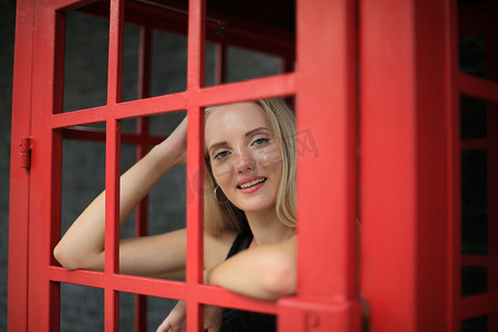 身穿黑色连衣裙的美丽金发女孩的画像站在红色电话亭里，靠在黑墙上，作为户外肖像时尚姿势。