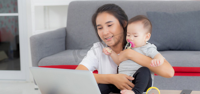 父母和孩子视频摄影照片_年轻的亚洲母亲在笔记本电脑上工作，同时在家照顾女儿，妈妈和女婴坐在一起使用笔记本视频通话、父母和亲密关系、室内、家庭和商业概念。