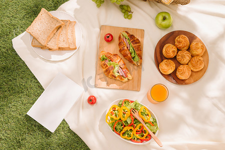 绿色格子布摄影照片_野餐柳条篮，里面有食物、面包、水果和橙汁，放在红色和白色的格子布上，背景是绿色。