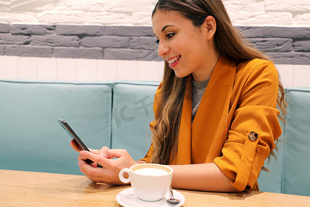 咖啡馆城市生活方式女孩坐在时尚的城市咖啡馆里，在智能手机应用程序上打电话喝卡布奇诺发短信
