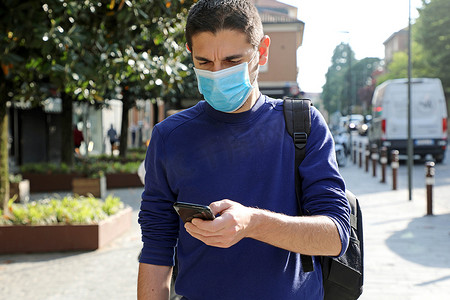 智能诊断摄影照片_COVID-19 大流行冠状病毒担心戴外科口罩的年轻人在城市街道使用智能手机应用程序帮助接触者追踪和自我诊断以应对 2019 年冠状病毒大流行