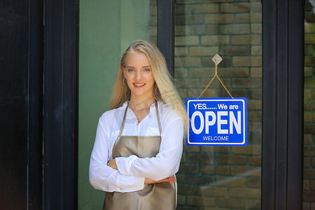 咖啡店标志摄影照片_自信的年轻女性商店的肖像，小企业咖啡店的老板站在店前，上面有开放的标志。