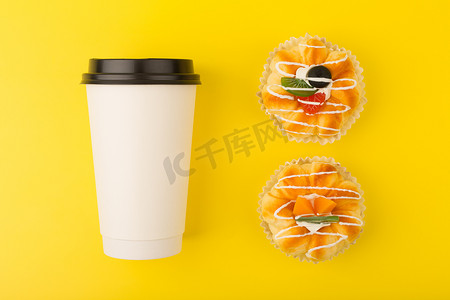 茶杯蛋糕摄影照片_白色纸板咖啡或茶杯和两个迷你蛋糕，黄色背景上有水果片