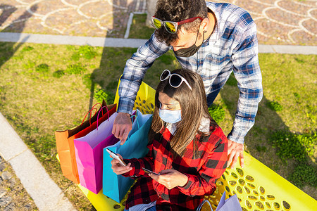 一对戴着防护面具的年轻夫妇坐在城市公园的长椅上，坐在许多彩色购物袋中，使用智能手机通过互联网移动连接查看在线购物交易。