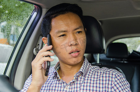 身穿格子衬衫的亚洲男子在车里看着智能手机。