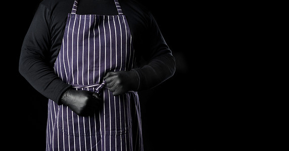 黑色衣服的人摄影照片_身穿蓝色条纹围裙和黑色衣服的男厨师站在黑色背景中