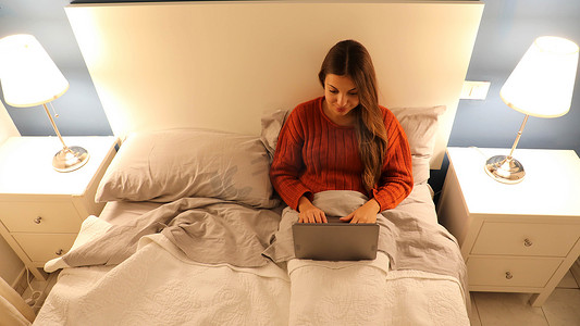 年轻美女夜间在双人床上使用笔记本电脑