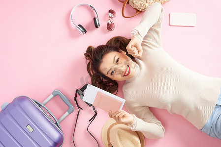 旅游行李卡片摄影照片_年轻的亚洲女性躺在粉红色的地板上，周围放着旅行行李，出示了她的护照。