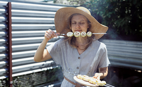 闻肉味道摄影照片_戴着夏日帽子的年轻女人在户外闻着烤蔬菜的味道