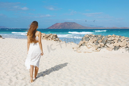 在加那利群岛富埃特文图拉的科拉莱霍海滩上，美丽的女人在科拉莱霍海滩上散步，看着罗伯斯岛，沙滩上有石头庇护所，以抵御强风