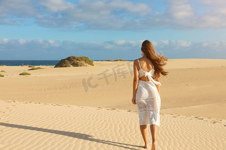 年轻漂亮的女人穿着白色的裙子在日落时分走在沙漠的沙丘上。