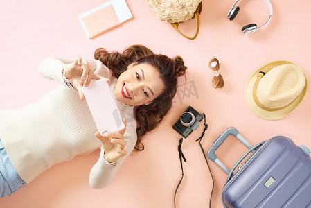 旅游行李卡片摄影照片_年轻的亚洲女性躺在粉红色的地板上，周围放着旅行行李，展示着她的智能手机。