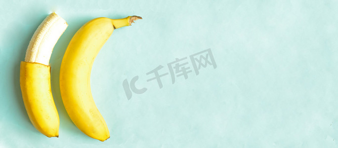 炎热夏季摄影照片_一根部分去皮的香蕉和一根完整的蓝色背景，复制空间。炎热夏季的甜热带水果。