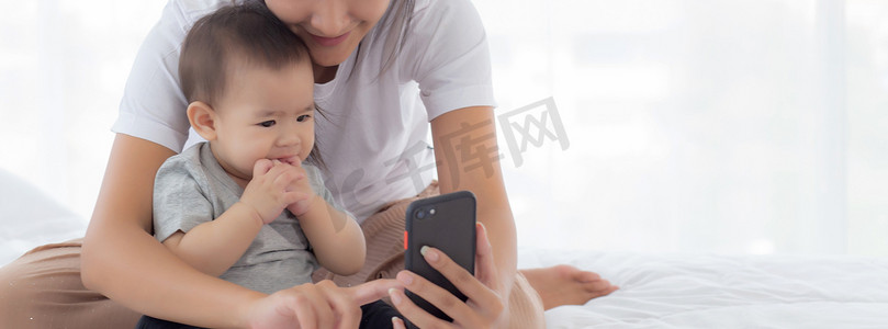 周年预热视频摄影照片_年轻的亚洲母亲和小女婴或新生儿在卧室床上用智能手机自拍，幸福的妈妈和女儿在家里使用电话视频通话，两个人，家庭和沟通概念。