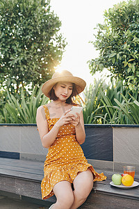 年轻的亚洲女孩在公园里使用智能手机，穿着带帽子的黄色连衣裙。