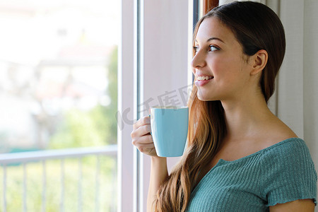 美女透过窗户在家喝茶的特写，关注模特的眼睛，室内照片