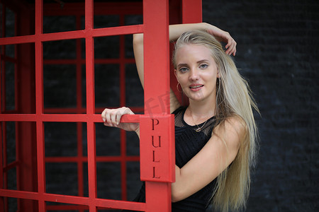 身穿黑色连衣裙的美丽金发女孩的画像站在红色电话亭里，靠在黑墙上，作为户外肖像时尚姿势。