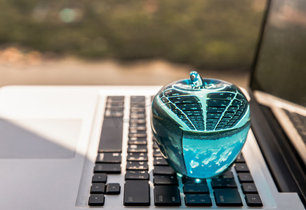 在阳台和自然景观的木桌上的笔记本电脑键盘上的玻璃水晶蓝苹果。