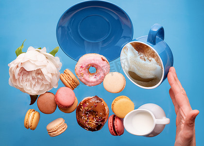 牡丹、蓝色杯子里的咖啡帽、甜甜圈、甜甜圈、马卡龙、牡丹和蓝色背景上飞舞的手持碟