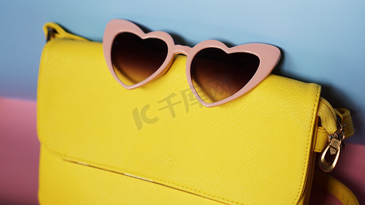 粉色夏天背景摄影照片_粉色和蓝色背景中的黄色包和心形太阳镜