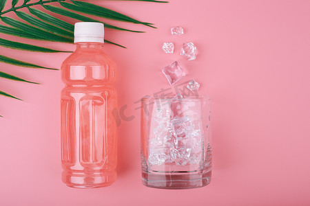 创意瓶中摄影照片_创意夏日组合物，瓶中有粉红色柠檬水，粉红色背景上有洒冰的玻璃杯，有棕榈叶