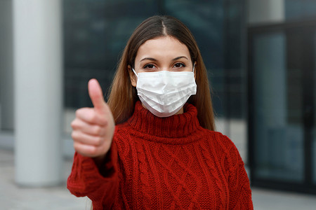 COVID-19 戴着防护面具的乐观年轻女性在城市街道上竖起大拇指，准备参加 2019 年冠状病毒病疫苗接种活动（Sars-Cov-2）