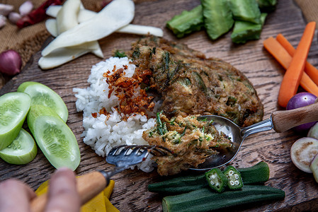 调cha摄影照片_泰国当地的美食风格，手用勺子在茉莉花米饭上舀油炸金合欢煎蛋卷或 cha-om 鸡蛋，配料和木制背景上的新鲜蔬菜，