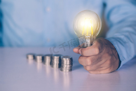 投资储蓄摄影照片_商人的手拿着灯泡，同时在办公桌上投币，为成功储蓄和融资，关于存款和收入和投资的知识，解决方案和灵感，商业理念。