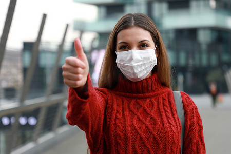 戴着防护面具的乐观年轻女性在城市街道上竖起大拇指，准备参加 2019 年冠状病毒病疫苗接种活动