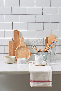 白色木墙上的简单质朴厨具：带木制炊具套装的粗糙陶瓷锅、成堆的陶瓷碗、水壶和木托盘。