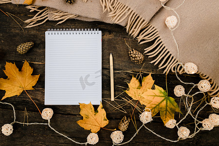 深色背景中的白色空白笔记本和笔，周围有围巾、格子花呢和杯子、秋天的黄叶和松果。