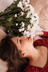 床上躺着一束雏菊，身穿红色裙子的漂亮褐发女郎