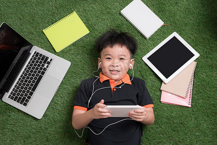 可爱的小男孩带着笔记本电脑、平板电脑和笔记本在草地上听音乐。