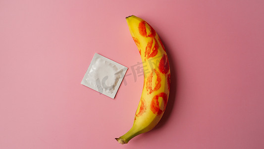 粉红色工作室背景中黄色香蕉上未包装的避孕套和红色唇膏