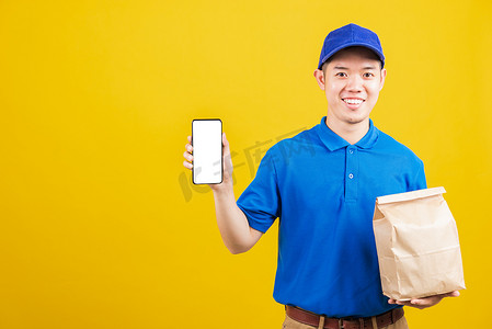 送货服务人员微笑着穿着蓝色制服拿着纸容器来装外卖袋杂货食品包并展示智能手机