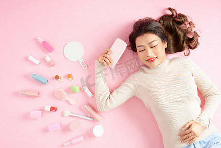 亚洲女人拿着她的手机，躺在粉红色的背景上，拿着她的美容工具。