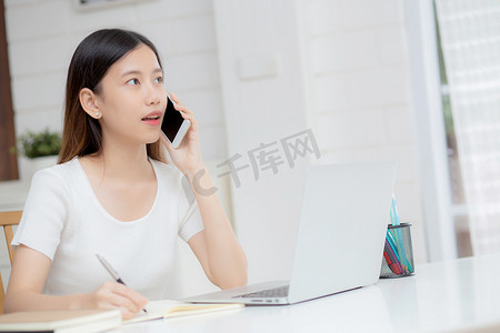 年轻的亚洲女性通过笔记本电脑在家打电话和工作，自由职业者在笔记本上写作并在智能手机上讲话，待在家里，家庭生活，商业和沟通理念。