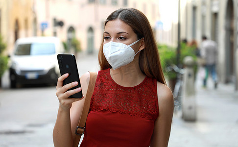 COVID-19 年轻女性佩戴 KN95 FFP2 面罩在城市街道使用智能手机应用程序以帮助接触者追踪和自我诊断以应对 2019 年冠状病毒病的肖像
