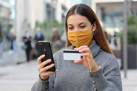 戴着防护面罩的年轻美女肖像在街上通过信用卡和智能手机应用程序冷静下来并付款