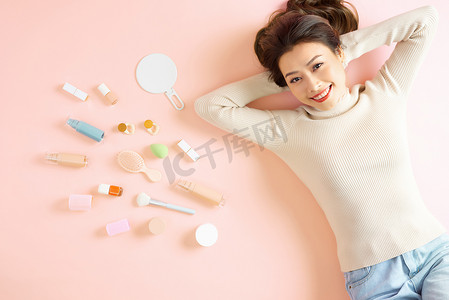 拿口红摄影照片_漂亮的亚洲女人微笑着躺在粉红色的地板上，拿着她的化妆工具。